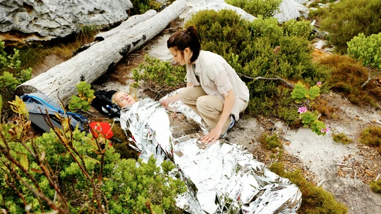 A woman lying on the ground wrapped in a shock blanket, while another woman crouches nearby assisting